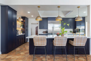 Cabinetry: Deep navy blue inset custom cabinets with gold hardware, offering a luxurious and modern aesthetic. Open shelving with warm lighting, creating a functional yet stylish area for displaying décor or everyday items. Countertop: A white quartz countertop on the island, combining sleek durability with elegance. The island provides ample workspace and includes a cooktop integrated into the design. Appliances: A large, high-end stainless steel refrigerator is the focal point of the back wall, seamlessly integrated into the layout. Built-in wall oven and microwave add to the functionality while maintaining the modern look. Lighting: Brass pendant lights with dome-shaped shades hang above the island, adding warmth and sophistication to the space. Recessed ceiling lights enhance brightness and keep the kitchen well-lit. Flooring: Hexagonal terracotta-style tiles add a touch of rustic charm, complementing the modern elements with a timeless feel. Seating: Wicker-style bar stools with metal frames surround the island, offering comfortable seating while contributing to the inviting atmosphere. Decorative Elements: A backsplash with a patterned tile design behind the coffee station, adding a touch of character and artistry. Plants and decorative items on the island and open shelving provide a fresh and vibrant feel. This kitchen blends modern design elements with warm, functional details, creating an inviting and stylish space perfect for family gatherings and entertaining