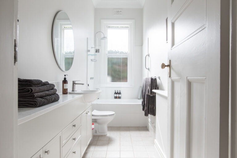 Small white bathroom with a vanity, round mirror, and bathtub under a bright window