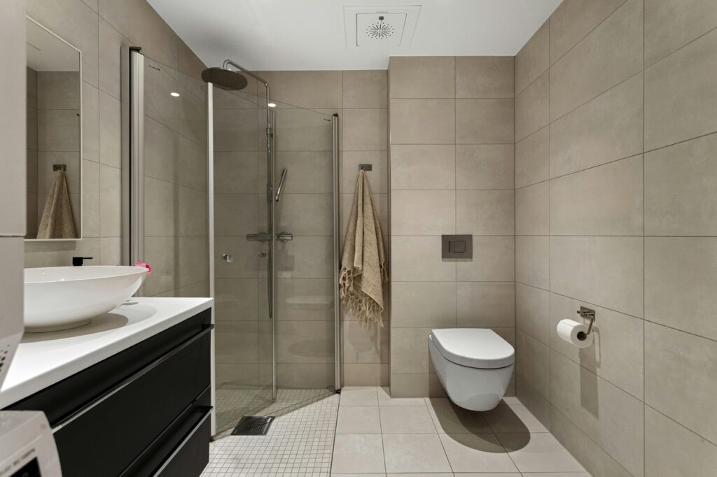 Modern bathroom with large beige tile flooring, glass walk-in shower, and wall-mounted toilet — ideal for clean, low-maintenance remodels