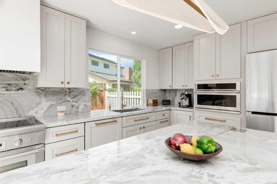 A photograph of a modern kitchen featuring light grey shaker-style cabinets, white marble countertops with grey veining, brass hardware, and stainless steel appliances, including a built-in oven and microwave. A large island with a bowl of fruit sits in the foreground, and a window looking out onto a backyard is above the sink.